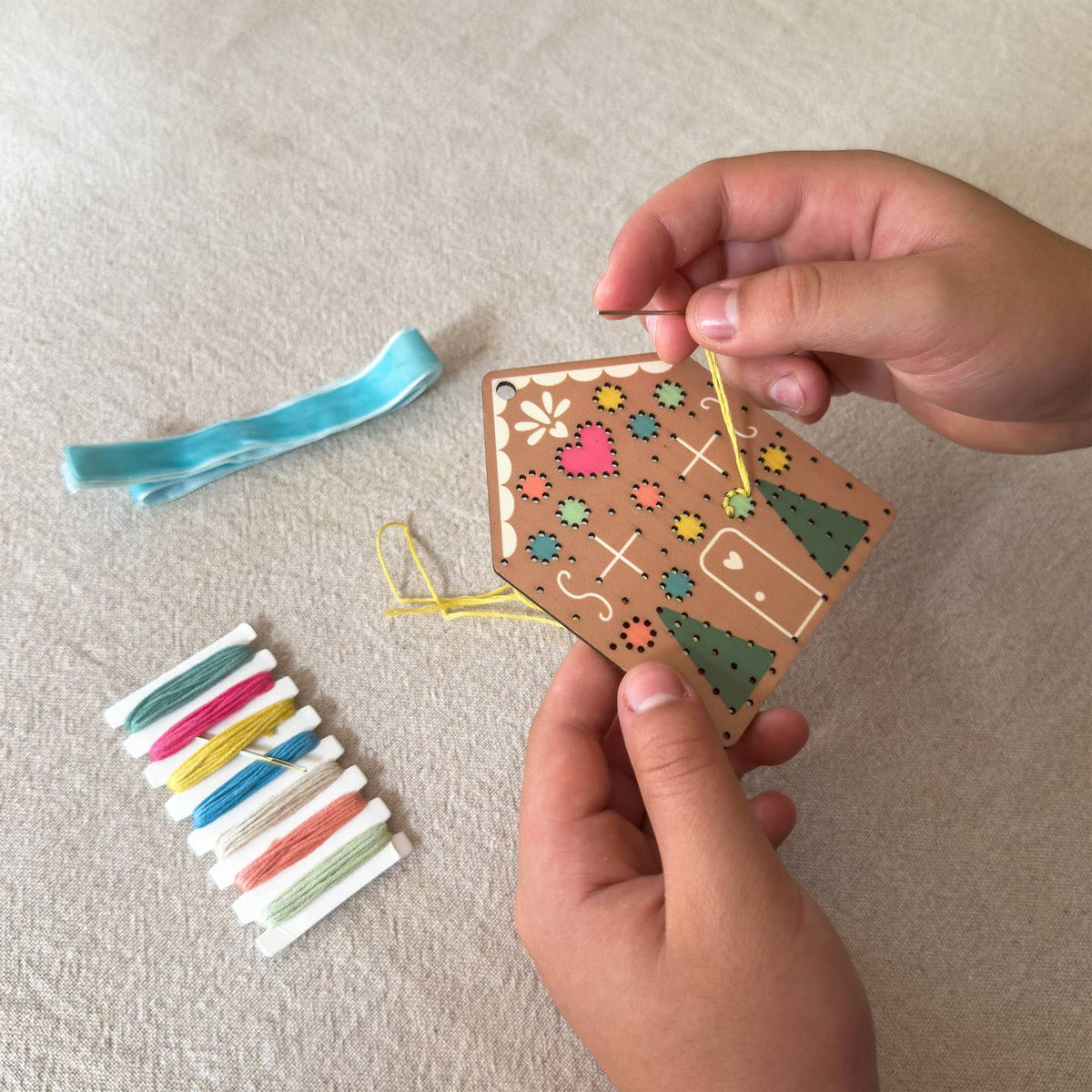 A person's hands are shown holding a wooden house-shaped embroidery kit over a beige fabric surface, with a blue ribbon and a small spool of multicolored thread nearby.