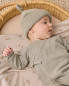 A baby lying on a patterned blanket inside a woven bassinet, wearing a textured sage green knit hat with a small pom-pom and a matching sage green long-sleeve romper.