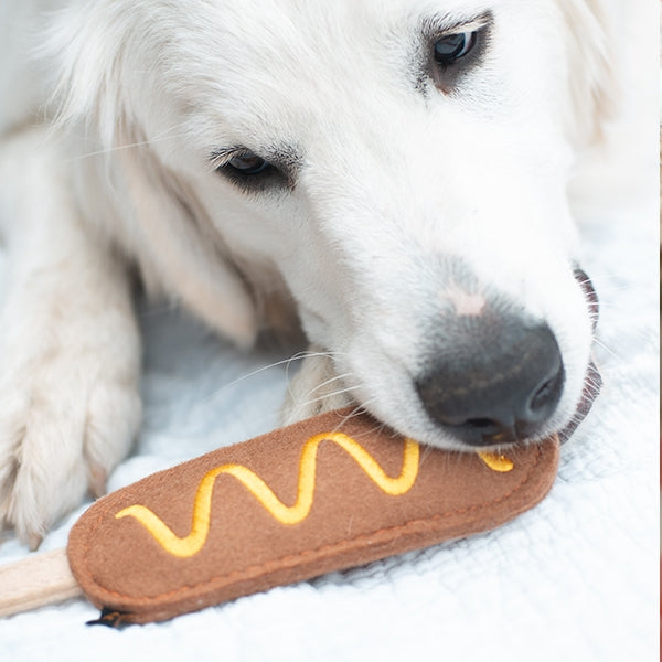 A white dog lying on a white blanket, chewing on a brown felt hot dog toy with yellow wavy lines.