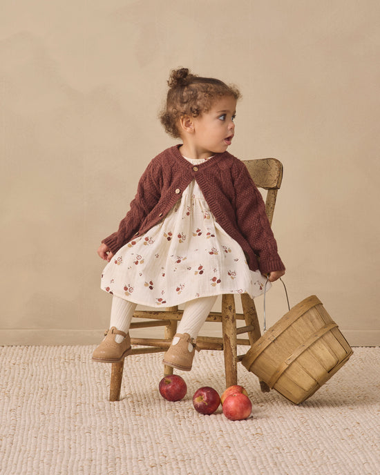 A toddler girl sits on a small wooden chair in a studio setting, wearing a dark brown knit cardigan over a cream floral dress with white socks and tan shoes. A wooden basket and a few red apples are on the floor beside her against a warm beige backdrop.
