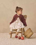 A toddler girl sits on a small wooden chair in a studio setting, wearing a dark brown knit cardigan over a cream floral dress with white socks and tan shoes. A wooden basket and a few red apples are on the floor beside her against a warm beige backdrop.