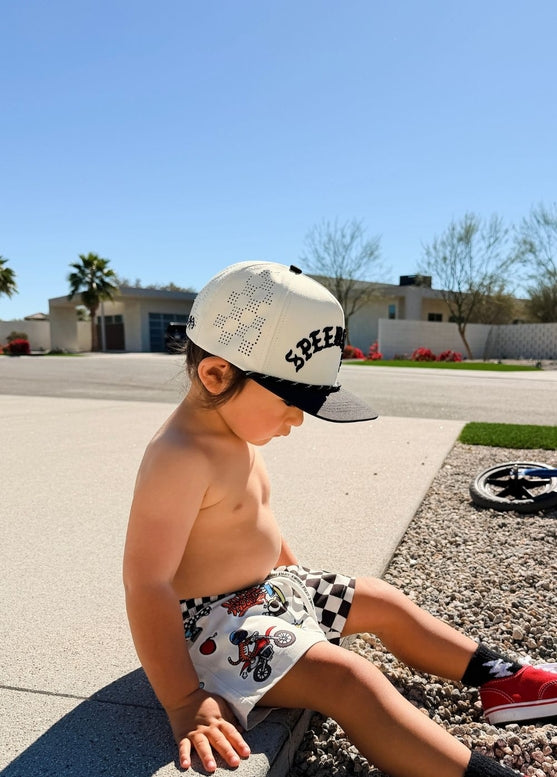 Child sitting on a driveway wearing a cap and checkered shorts.