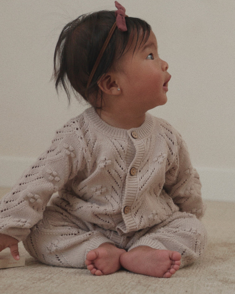 A baby sitting on a beige carpet wearing a matching beige knit cardigan and pants set with a pink bow headband.