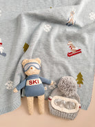 A cozy knit blanket in pale blue-gray with embroidered ski motifs, shown styled with a plush teddy bear wearing a blue ski outfit and a gray knit pom-pom beanie with a clear window.