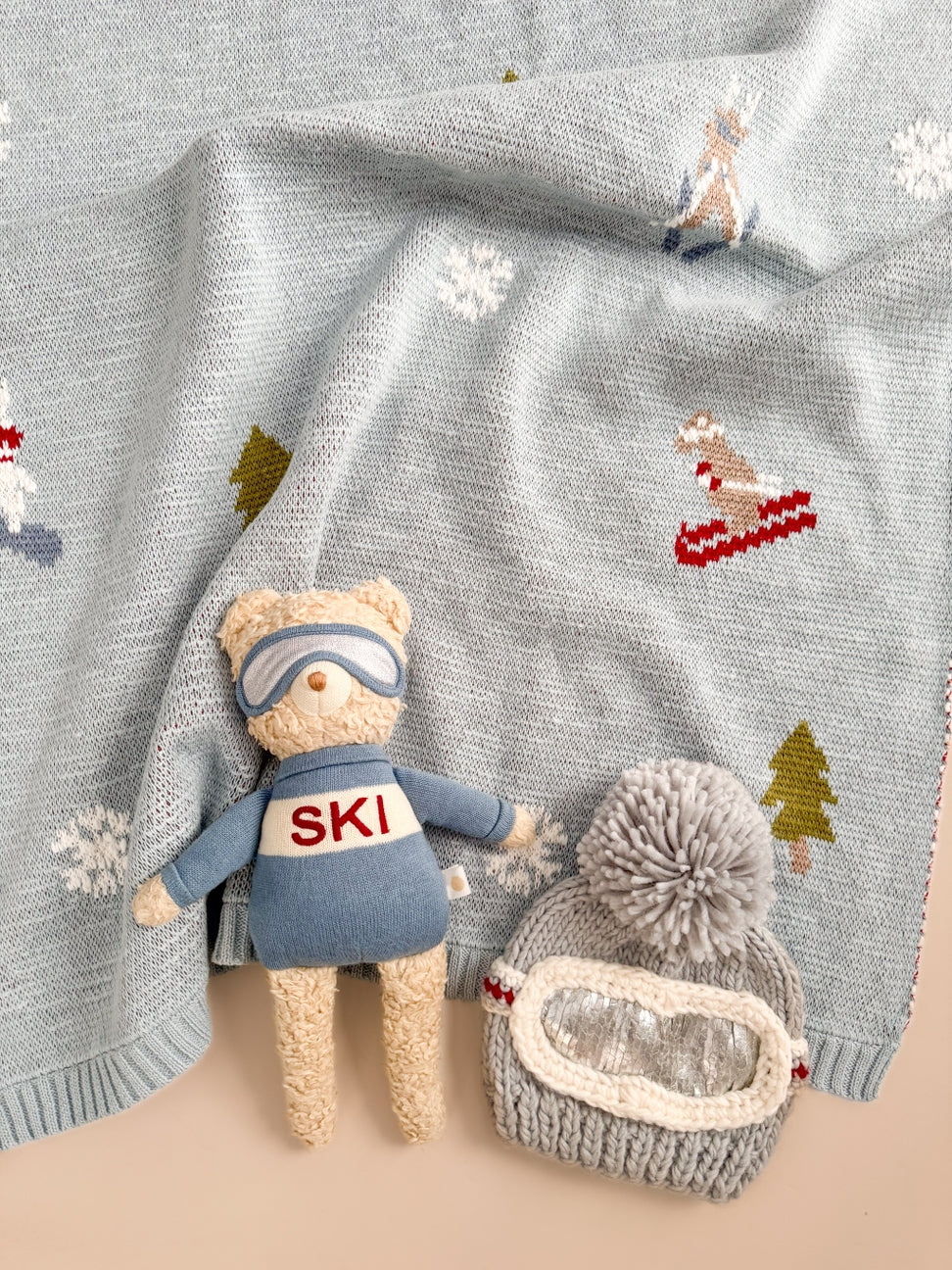 A cozy knit blanket in pale blue-gray with embroidered ski motifs, shown styled with a plush teddy bear wearing a blue ski outfit and a gray knit pom-pom beanie with a clear window.