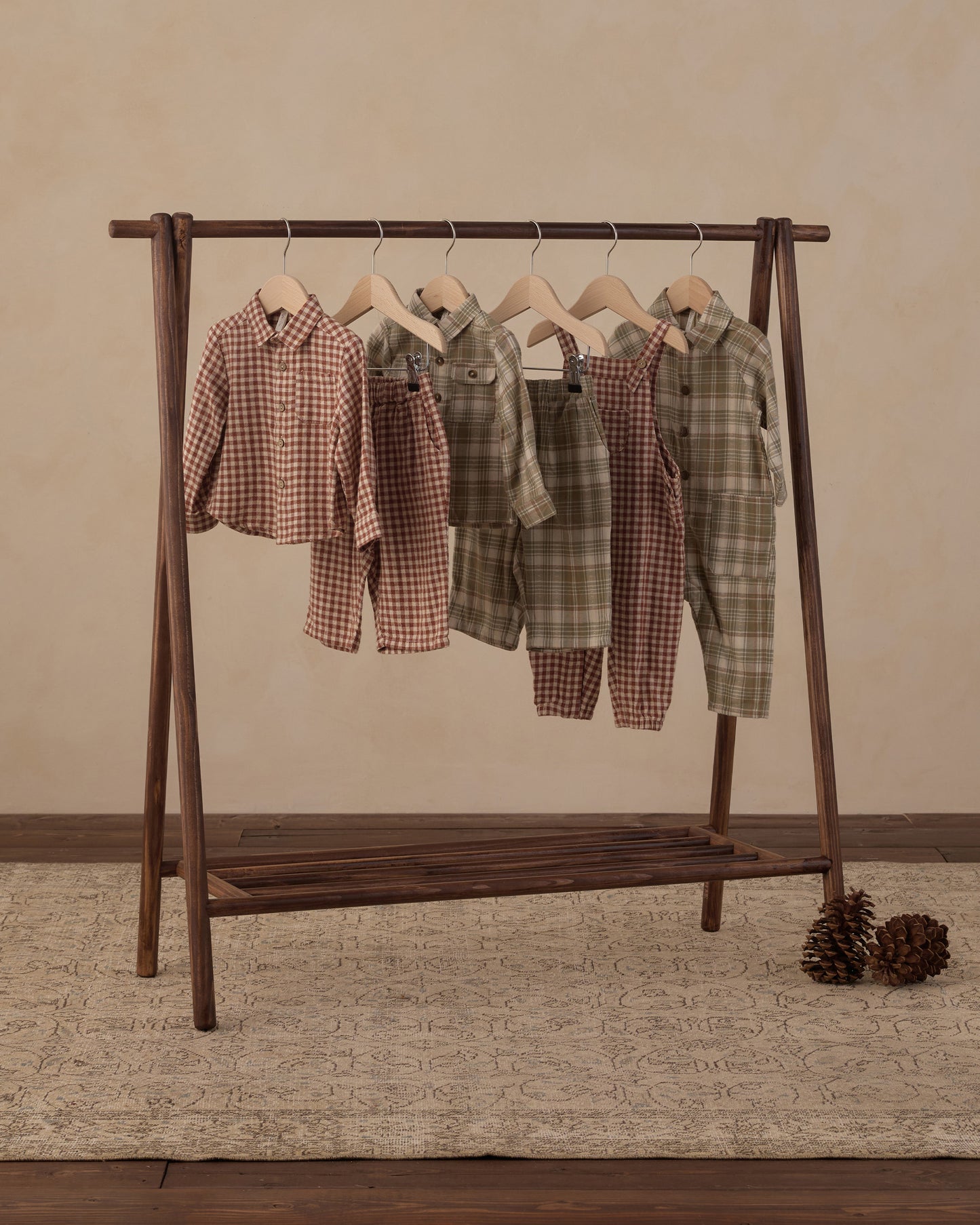 Five children's two-piece pajama sets in plaid and gingham patterns hang on a dark wooden A-frame clothing rack. The sets feature earthy tones including brown, green, and beige.