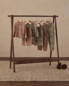 Five children's two-piece pajama sets in plaid and gingham patterns hang on a dark wooden A-frame clothing rack. The sets feature earthy tones including brown, green, and beige.
