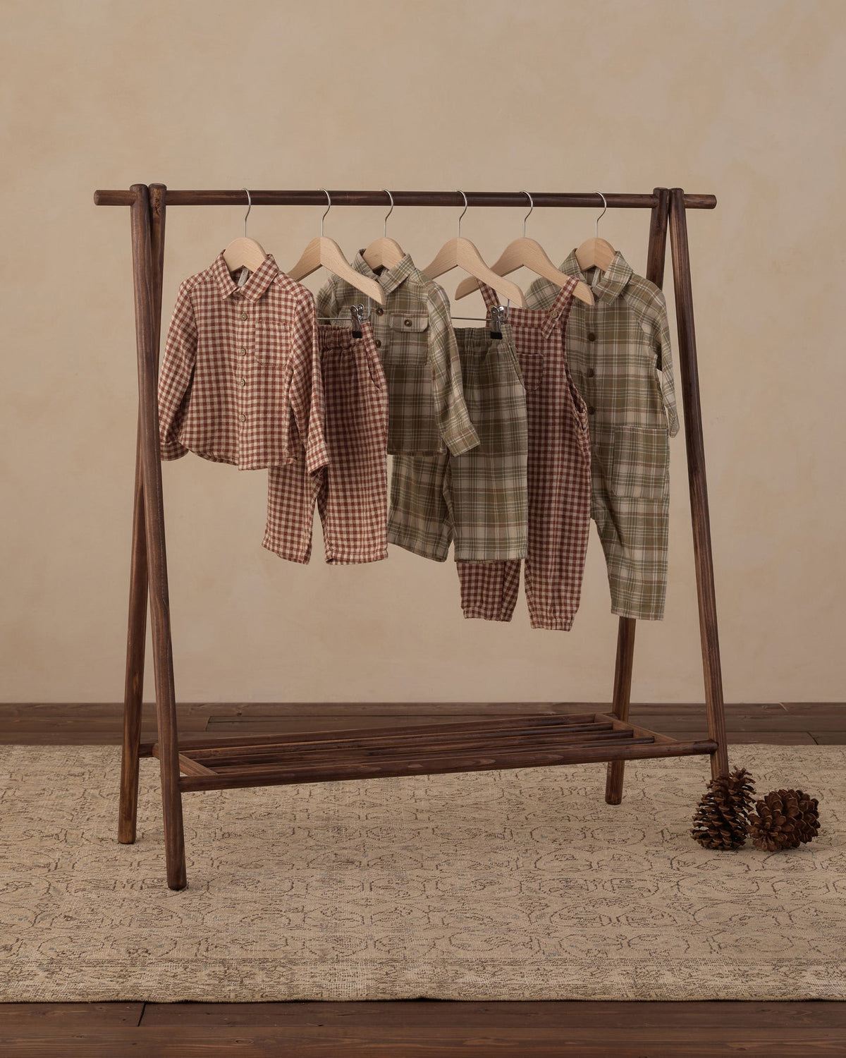 Five children's two-piece pajama sets in plaid and gingham patterns hang on a dark wooden A-frame clothing rack. The sets feature earthy tones including brown, green, and beige.
