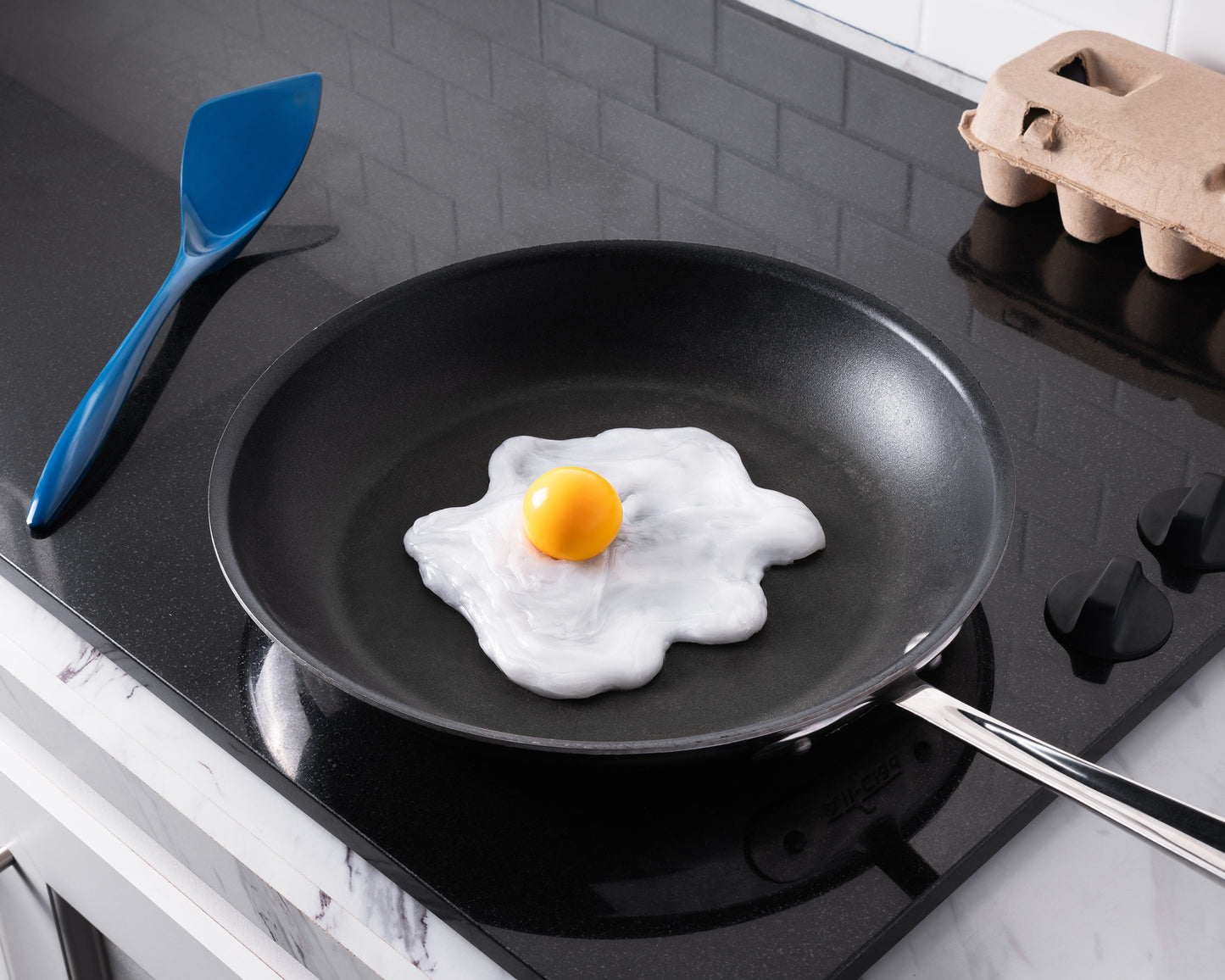 Fried egg in a black frying pan on a stovetop with a blue spatula and cardboard egg carton.