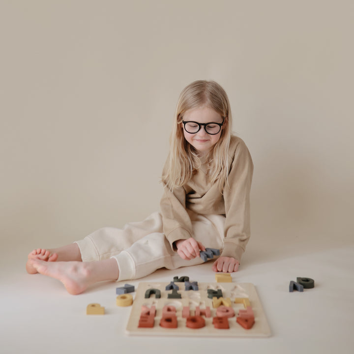 A young girl wearing glasses and a beige outfit sitting on the floor indoors, playing with wooden alphabet puzzle pieces scattered near a wooden puzzle board.