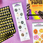 A styled flat-lay of Halloween-themed sticker products on a purple background. The products include a sheet of black alphabet stickers with yellow letters, a sheet of white ghost stickers with colorful faces, and a sheet of autumn pumpkin and face stickers.