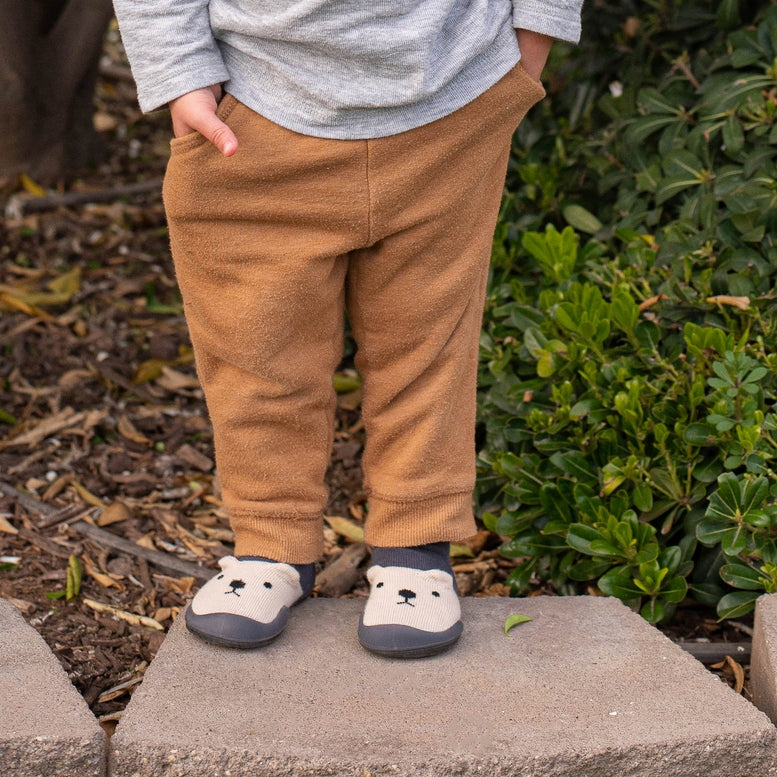 Child wearing brown pants and bear-themed shoes standing on a stone block with greenery in the background.