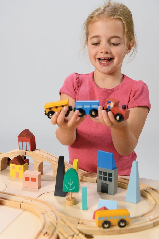 A young child with blonde hair wearing a pink shirt holds up a blue toy train engine and yellow cargo car, smiling playfully above a wooden toy train set and colorful town blocks.
