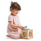 A toddler in a light pink dress sits on a plain white background, playing with a wooden activity cube by Mentari that features a colorful xylophone on top, spinning panels, and cogs on the side.