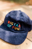 Navy blue cap with 'BIG FAN OF FUN' text in colorful letters on a wooden surface.