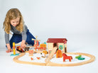 A young child lying on the floor, reaching out to play with a wooden farmyard-themed train set complete with tracks, a red barn, a windmill, and various animal figures.