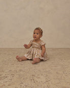 A baby girl sitting on a textured beige rug in front of a neutral beige backdrop, wearing a short-sleeve cream dress with a subtle floral pattern and a matching hair bow.