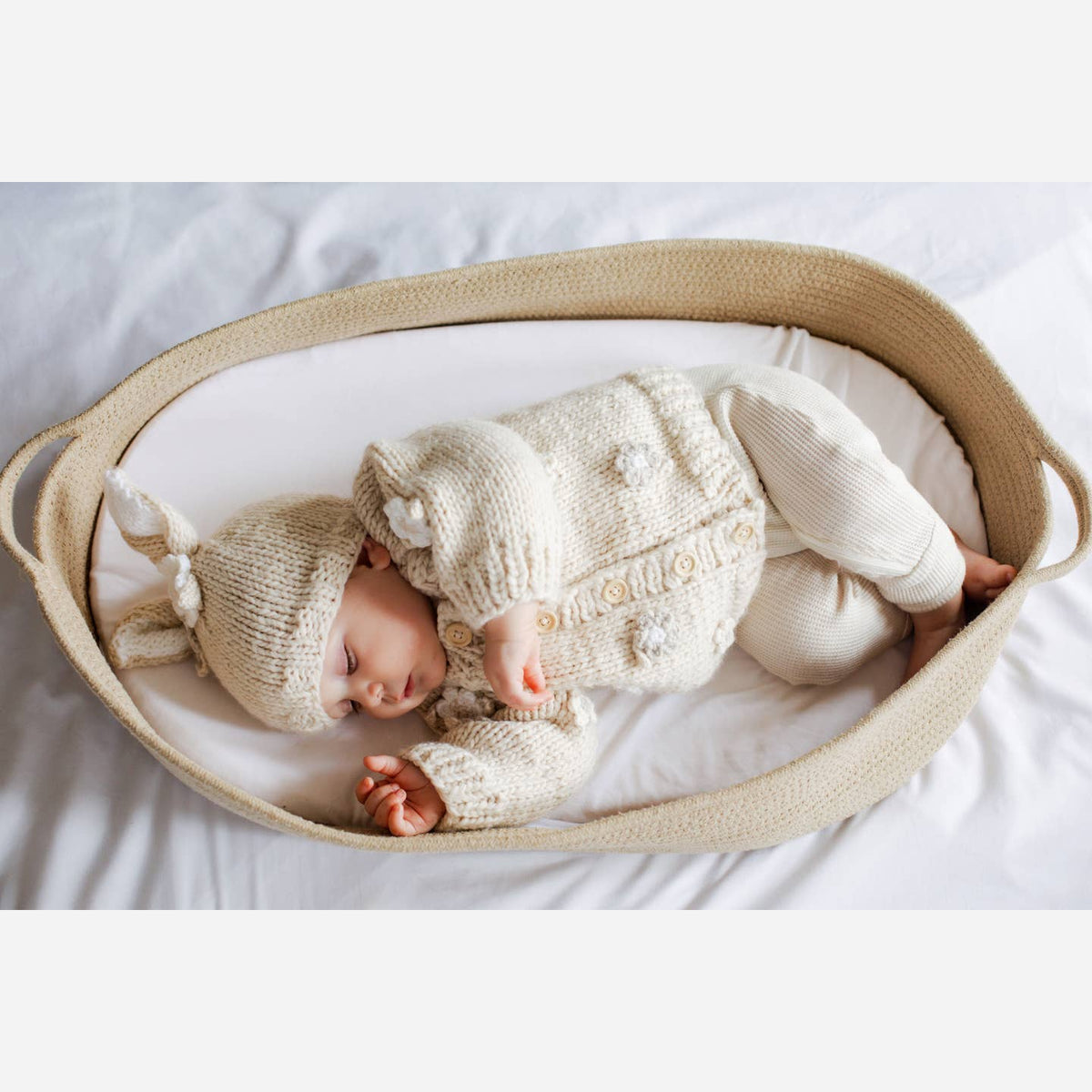 Baby sleeping in a bassinet wearing a knitted outfit and hat.
