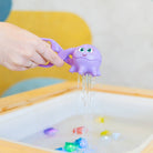 Purple octopus-shaped bath toy with water spout over a bathtub filled with water and colorful toys.