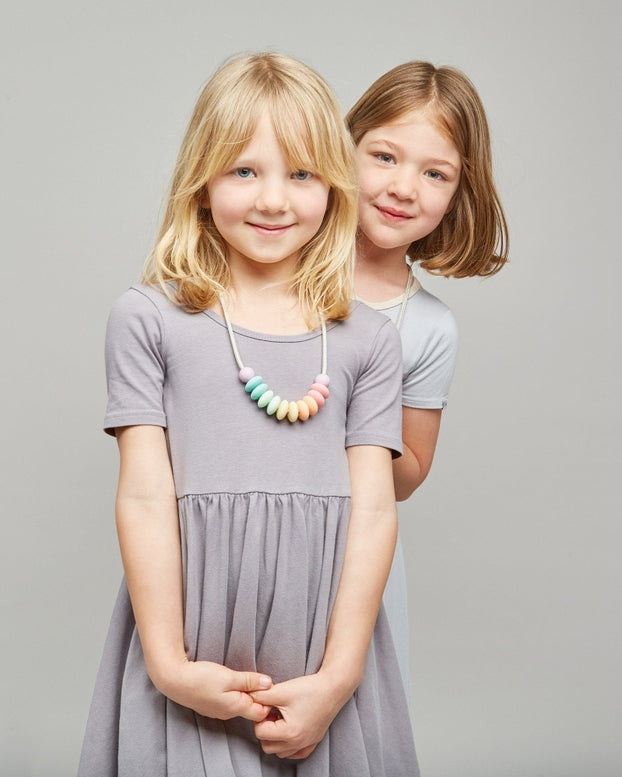 Two young girls modeling short-sleeve tiered dresses in a studio setting. The girl in the foreground wears a dark charcoal gray dress and poses with her arms crossed, while the girl behind her wears a light gray dress.