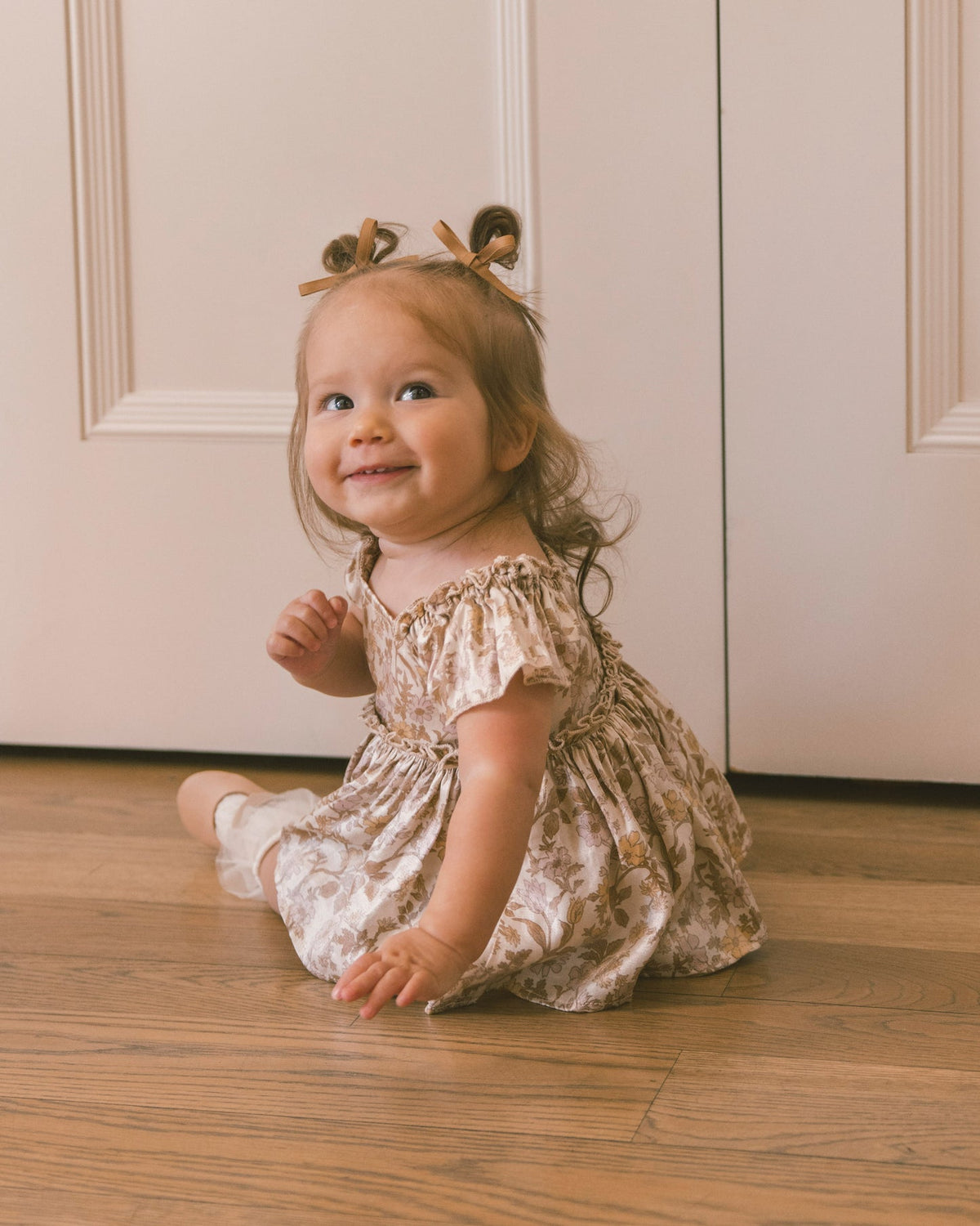 A toddler girl sitting on a wooden floor in front of cream-colored paneled doors, wearing a beige and brown floral print dress with a matching hair bow headband.