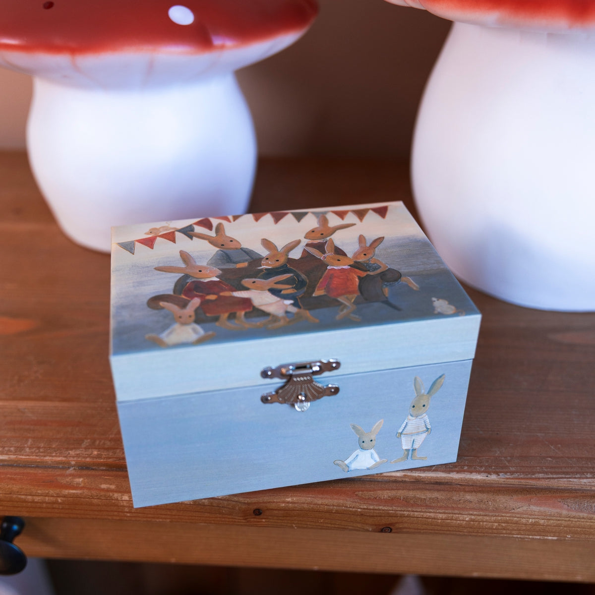 A small rectangular wooden box with a light blue front and lid, decorated with a painted scene of rabbits and two small rabbit figures, displayed on a wooden surface with mushroom-shaped decor in the background.