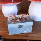 A small rectangular wooden box with a light blue front and lid, decorated with a painted scene of rabbits and two small rabbit figures, displayed on a wooden surface with mushroom-shaped decor in the background.