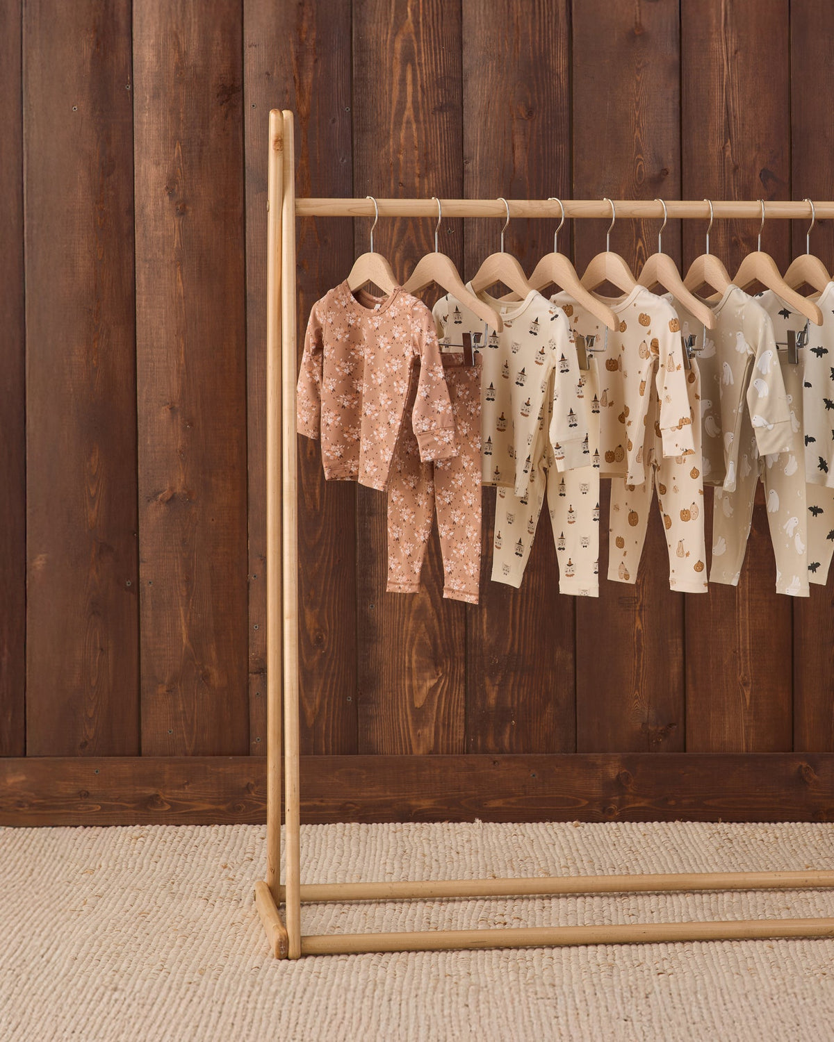 A wooden clothing rack holding several baby long-sleeve one-piece outfits in soft neutral tones, displayed against a dark wood plank wall and light woven rug.