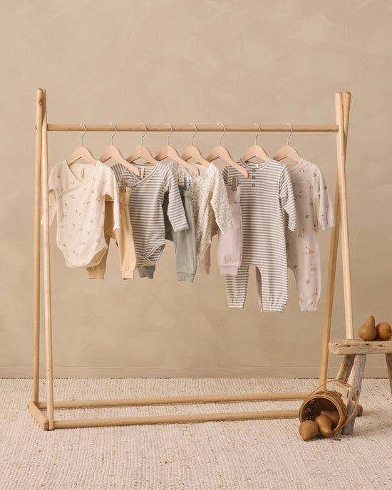 A set of baby bodysuits and long-sleeve tops in soft neutral tones, including stripes, solid creams, and light pinks, hanging on a light wood A-frame clothing rack. The scene is styled with a rustic wooden stool and pears on a textured beige rug against a warm beige wall.