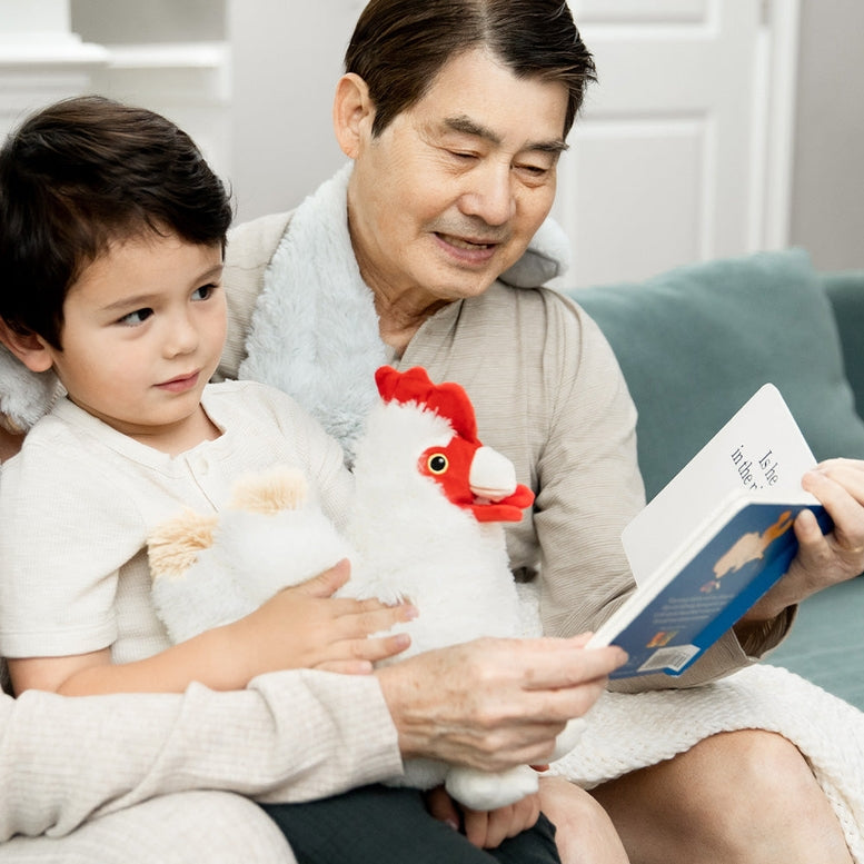 A young girl sitting on an adult's lap reading a book together while hugging a fluffy white Warmies chicken plush toy with a red comb and wattle.