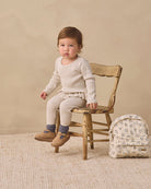 A toddler sits on a rustic wooden chair wearing a cream ribbed knit long-sleeve top and matching cream ribbed knit pants, with gray socks and tan shoes. A small cream floral backpack rests beside the chair against a warm beige studio backdrop.