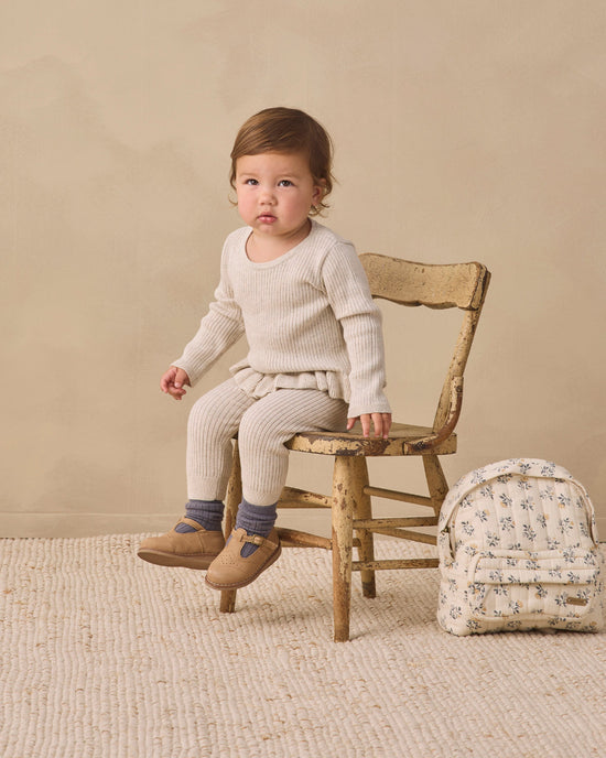 A toddler sits on a rustic wooden chair wearing a cream ribbed knit long-sleeve top and matching cream ribbed knit pants, with gray socks and tan shoes. A small cream floral backpack rests beside the chair against a warm beige studio backdrop.
