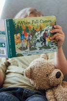 A toddler holding an open board book titled When Fall Comes, looking down at the colorful illustrated pages showing autumn trees and an outdoor nature scene, with a teddy bear resting nearby.