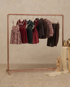 A copper-colored clothing rack displaying a collection of children's clothing, including plaid dresses, velvet dresses, a dark jacket, and trousers, set against a textured beige wall and floor.