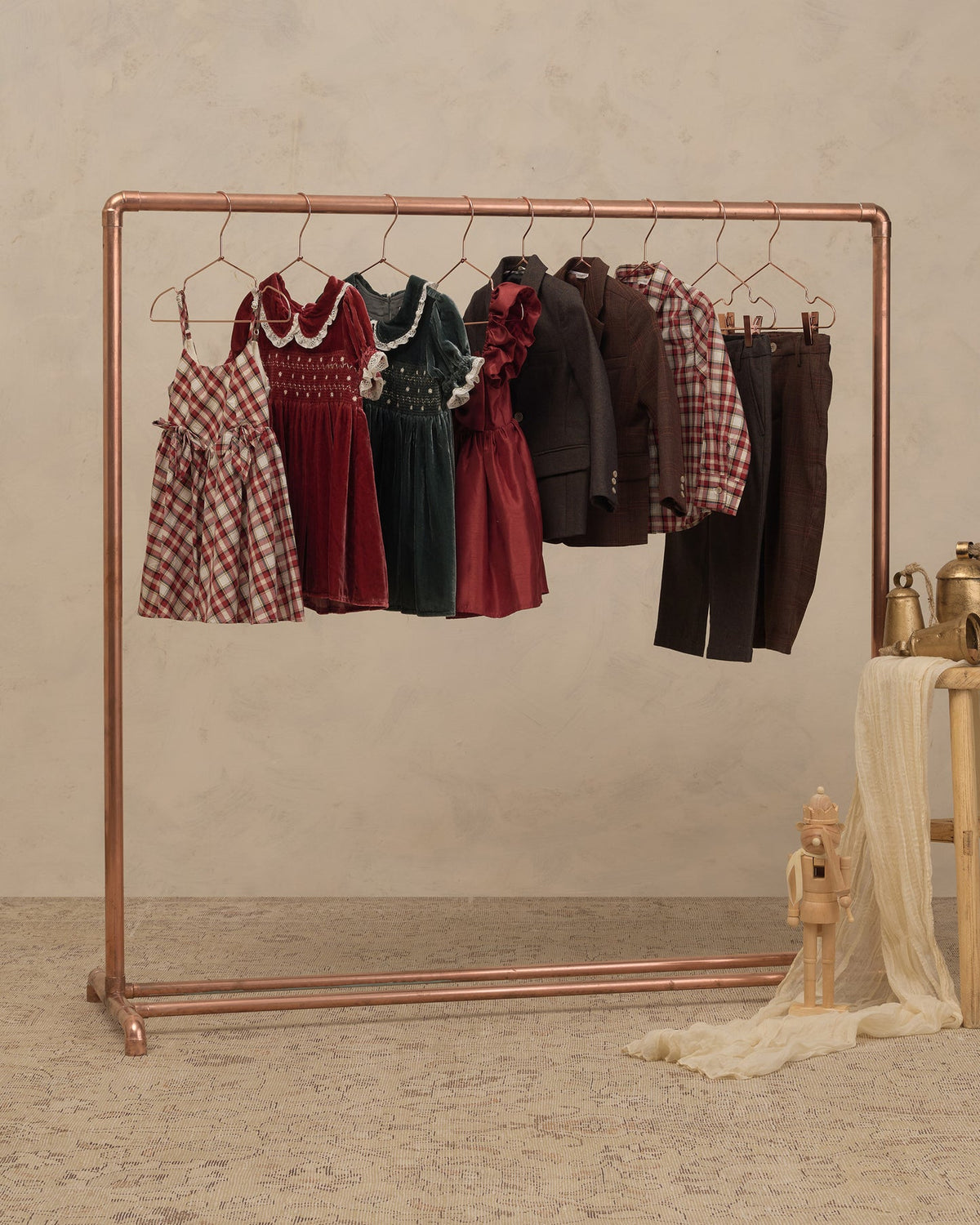 A copper-colored clothing rack displaying a collection of children's clothing, including plaid dresses, velvet dresses, a dark jacket, and trousers, set against a textured beige wall and floor.