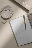 Notebook with a gold pen on a beige surface, with 'Cafés' text in the background.