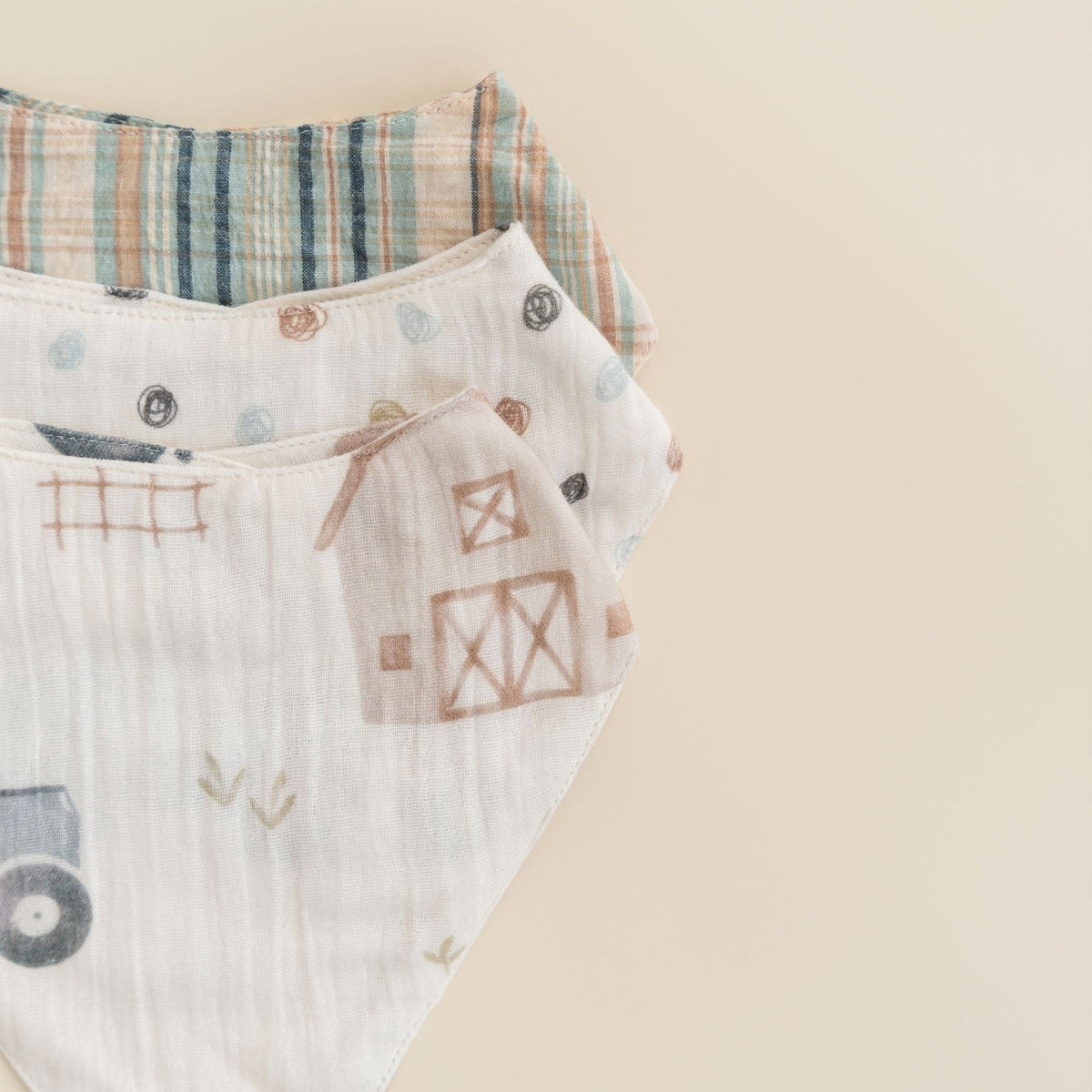 A close-up of a soft cotton muslin baby bandana or bib with a farm-themed print, featuring a barn, tractor, and plaid pattern on a warm beige background.