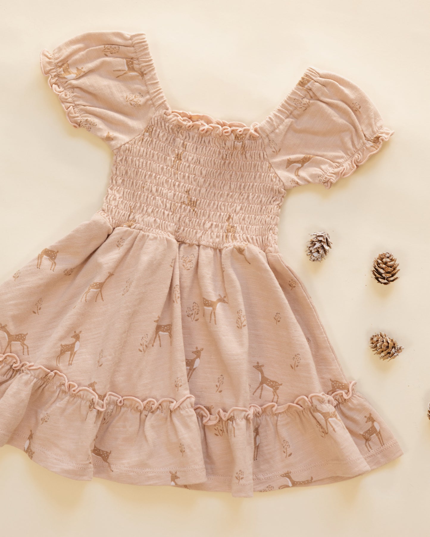 A baby girl's smocked dress in a dusty rose pink color, featuring a subtle deer print and ruffled trim, laid flat on a cream-colored background with decorative pinecones.