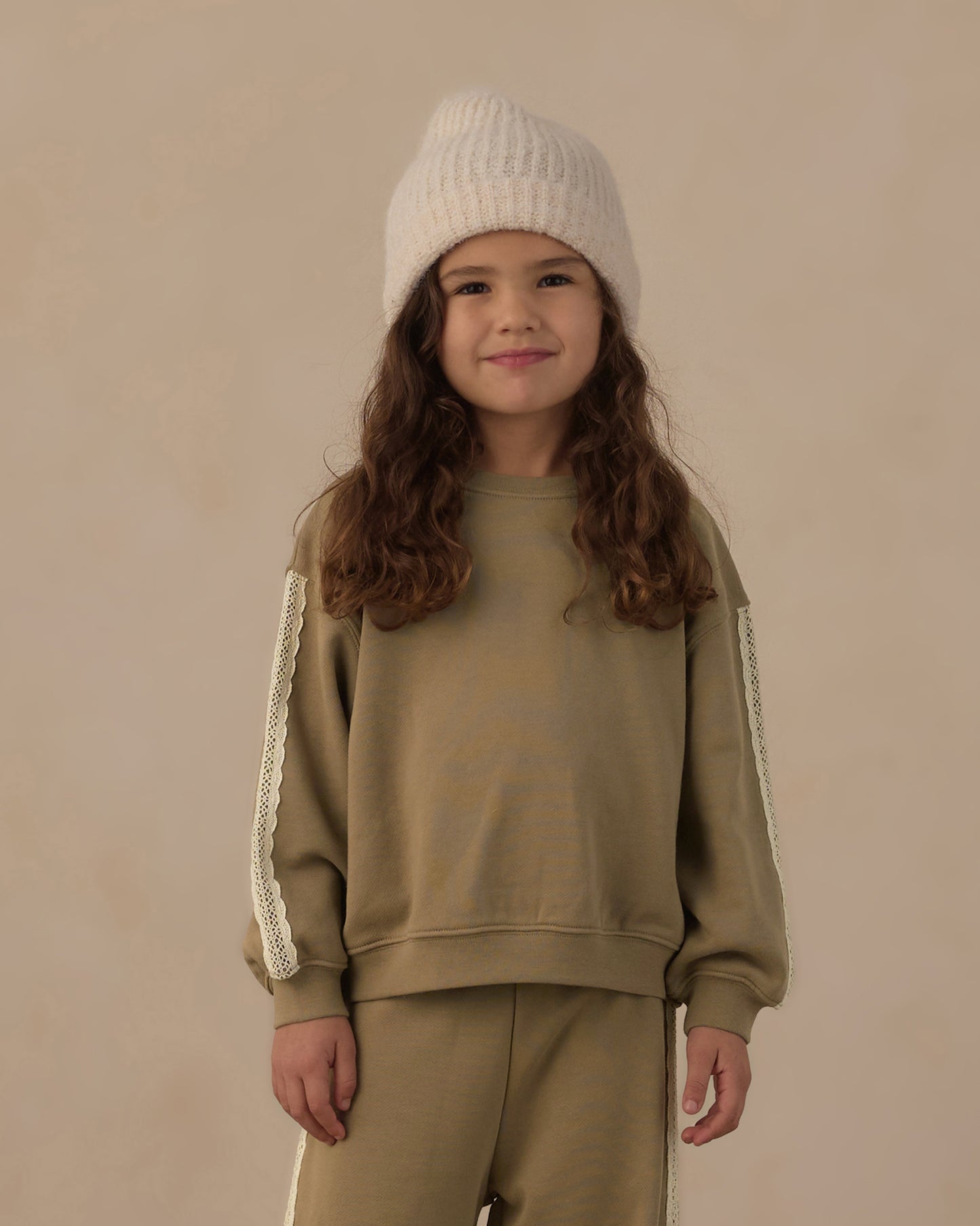A young girl with long wavy brown hair is wearing a matching beige sweatshirt and sweatpants set with cream lace sleeve details, paired with a cream knit beanie, against a solid beige background.