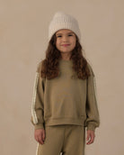 A young girl with long wavy brown hair is wearing a matching beige sweatshirt and sweatpants set with cream lace sleeve details, paired with a cream knit beanie, against a solid beige background.