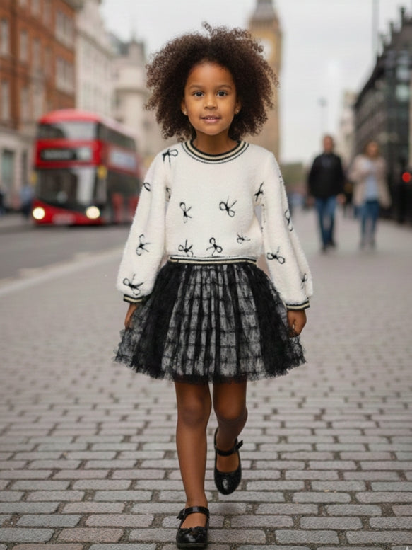 A young girl with curly dark hair walks on a cobblestone city street wearing a white knit sweater with black bow motifs and a black tulle skirt.