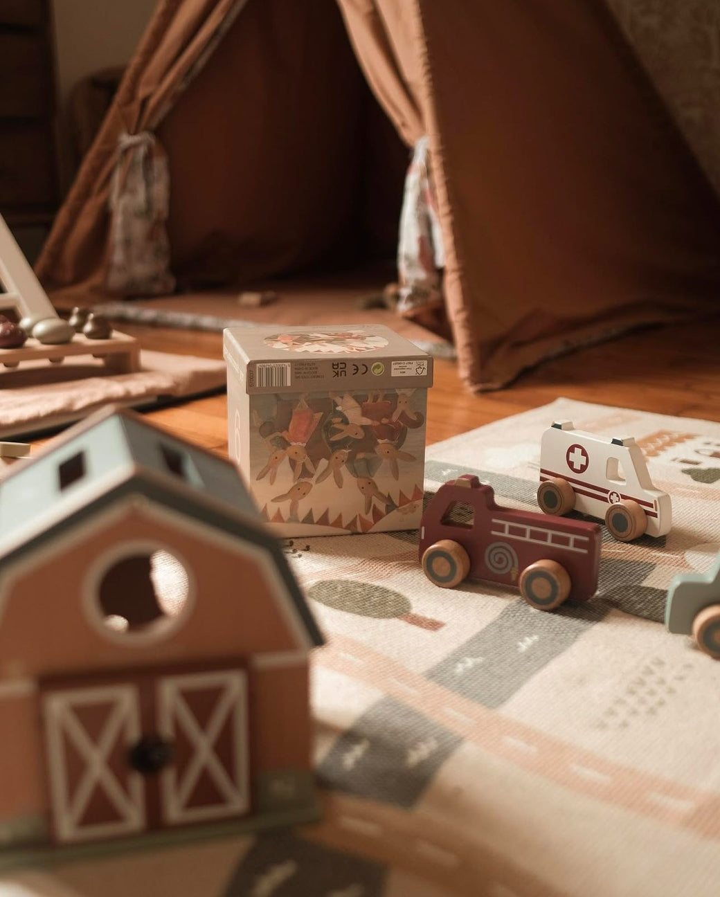 Children's toys including a wooden barn, fire truck, and ambulance on a wooden floor with a tent in the background.