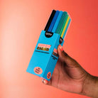 A hand holding a box of OMY Super Clean 12-Count Double Point Marker pens, showing the colorful tips of the markers through a clear plastic window on the box, set against a solid orange background.