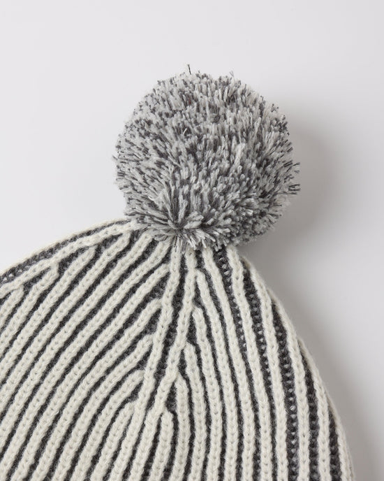 Close-up of a knit beanie with vertical black and off-white stripes and a fluffy gray pom-pom on top, photographed against a light gray background.