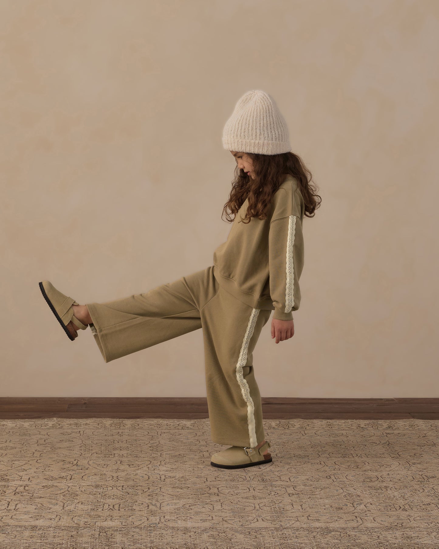 A child wearing a matching beige tracksuit with white side stripes, a cream knit beanie, and tan shoes, posing with one leg raised against a textured beige wall and patterned rug.