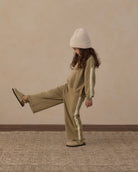 A child wearing a matching beige tracksuit with white side stripes, a cream knit beanie, and tan shoes, posing with one leg raised against a textured beige wall and patterned rug.