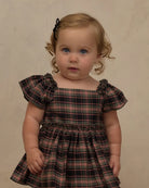 A toddler girl with blonde curly hair and blue eyes wearing a dark plaid dress with ruffled flutter sleeves, standing against a neutral beige studio backdrop.