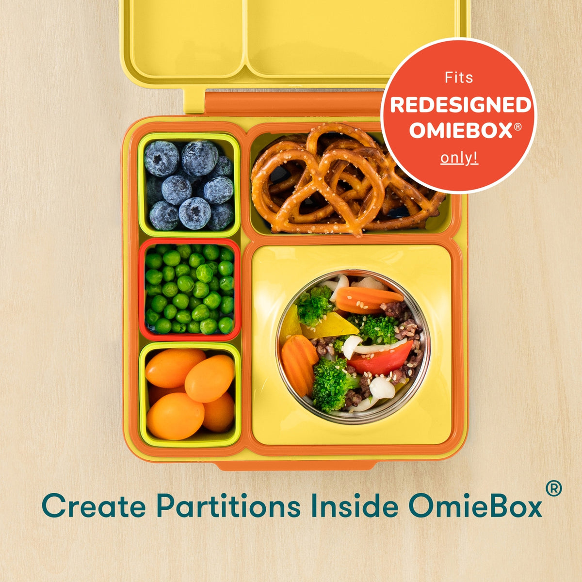 Top-down view of a yellow and orange OmieBox lunch container with divided compartments, shown on a light wood background. The container is filled with blueberries, pretzels, peas, carrots, and a small salad bowl.