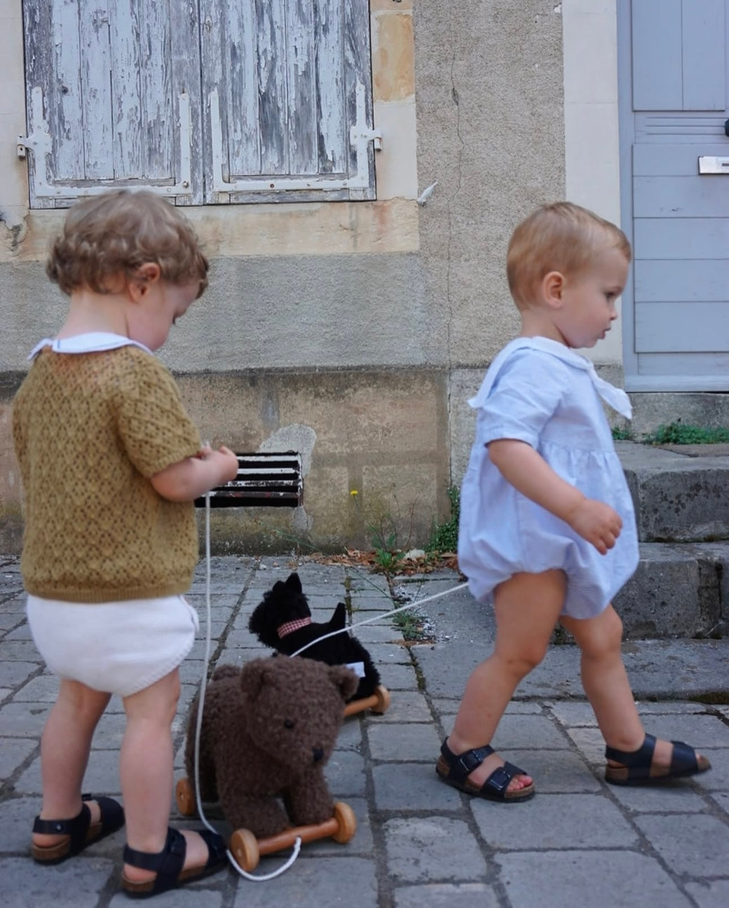 Two children playing on a stone pavement with a teddy bear and a small black dog toy.