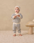 A baby standing barefoot in a studio setting, wearing a cream and gray striped knit long-sleeve top, matching pants, and a knit beanie. The background is a warm beige wall with a small wooden mushroom prop on the right.